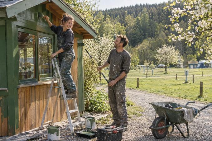 Frau auf einer Leiter und Mann mit Farbroller streichen im Frühling ein grünes Rezeptionshäuschen aus Holz, dahinter leere Stellplätze mit Stromsäulen
