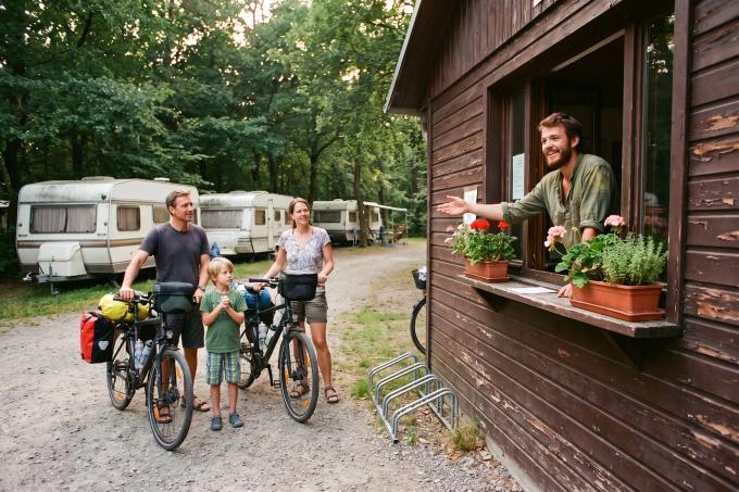 Ein junger Mitarbeiter begrüßt am Rezeptionsfenster eine Familie mit Fahrrädern.